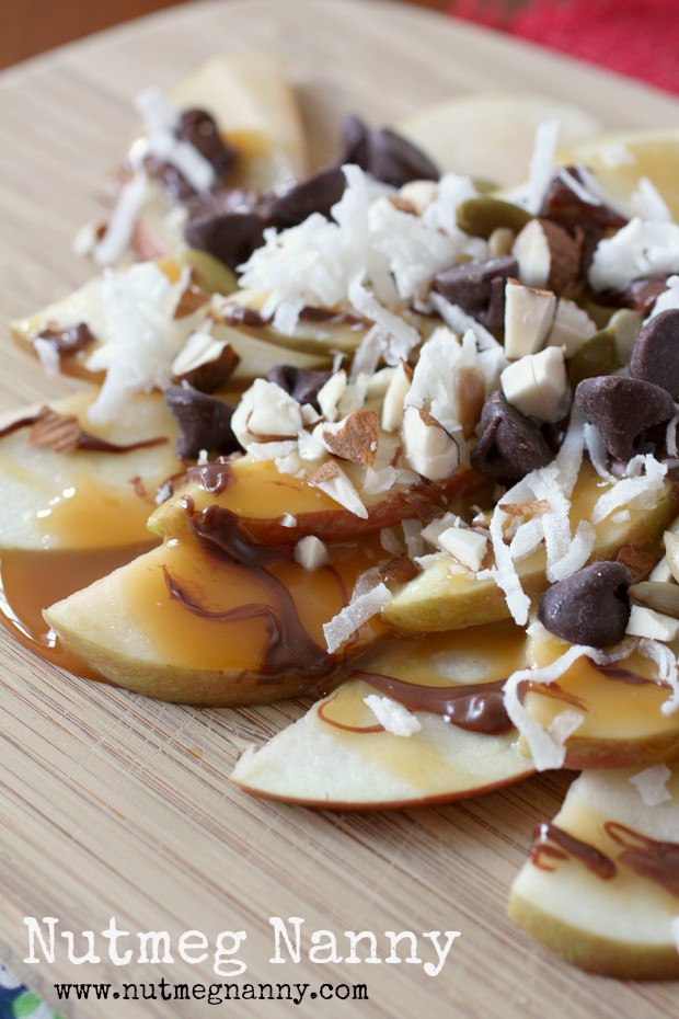Apple Nachos by Nutmeg Nanny apple nachos on a wooden plate topped with nuts, coconut and chocolate.
