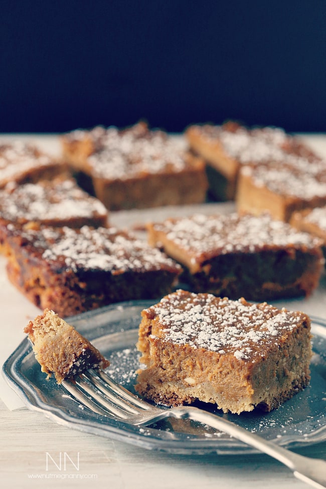 These pumpkin pie cake bars are like two desserts in one. Cake like bottom topped with a moist pumpkin pie top sprinkled with powdered sugar. Perfect for Thanksgiving! Pumpkin Pie Cake Bars on a plate with sliced bars behind it.