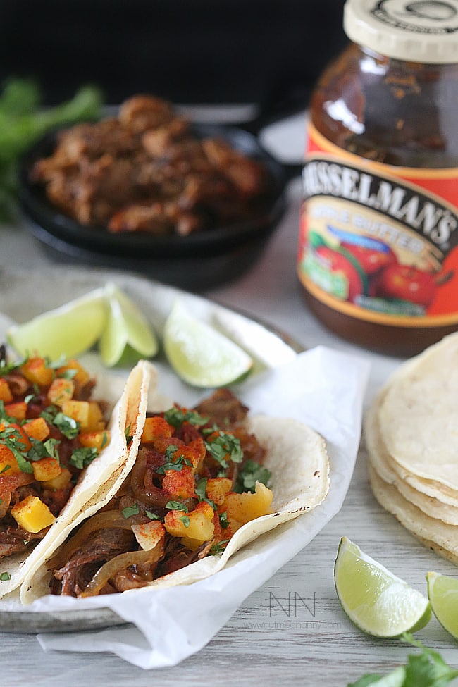 Carnitas in tortillas on a plate with lime wedges on the side and a jar of apple butter in the background.