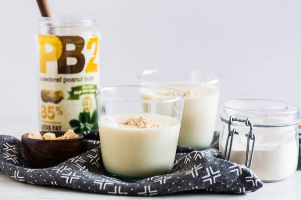 Two glasses of peanut butter protein shake on a table with containers of peanut butter powder and protein powder.