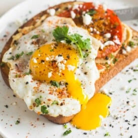 This shakshuka hummus toast is a healthy and hearty breakfast. Hummus spread toast topped with spiced roasted tomatoes, fried egg, cilantro and feta cheese.