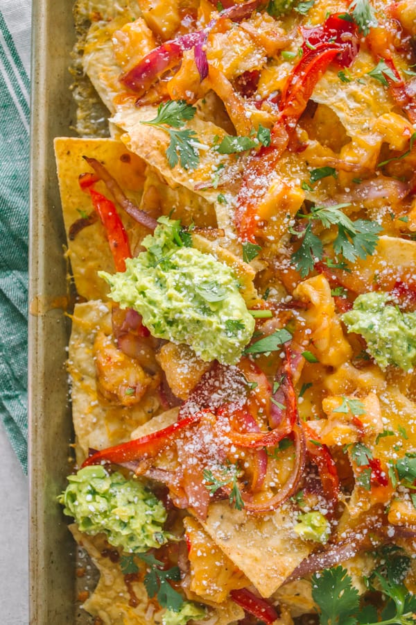 Close up of nachos on a sheet pan covered in chicken, cheese, onions, guacamole and cilantro.