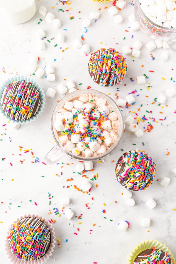 chocolate strawberry funfetti hot chocolate bombs on a white table with a mug of hot chocolate