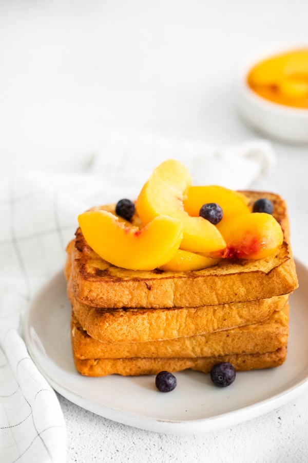 Peach Topped French Toast stacked on a plate topped with peaches and blueberries.