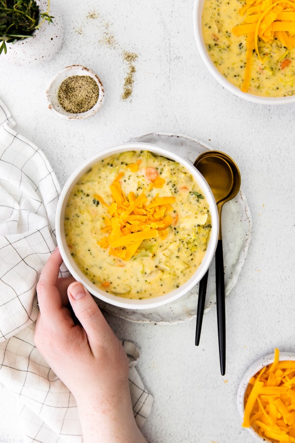 Instant Pot Broccoli Cheddar Soup in a bowl with a hand touching the side of the bowl. 