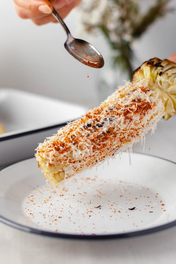 A hand sprinkling on chili powder to a cob of elote. 