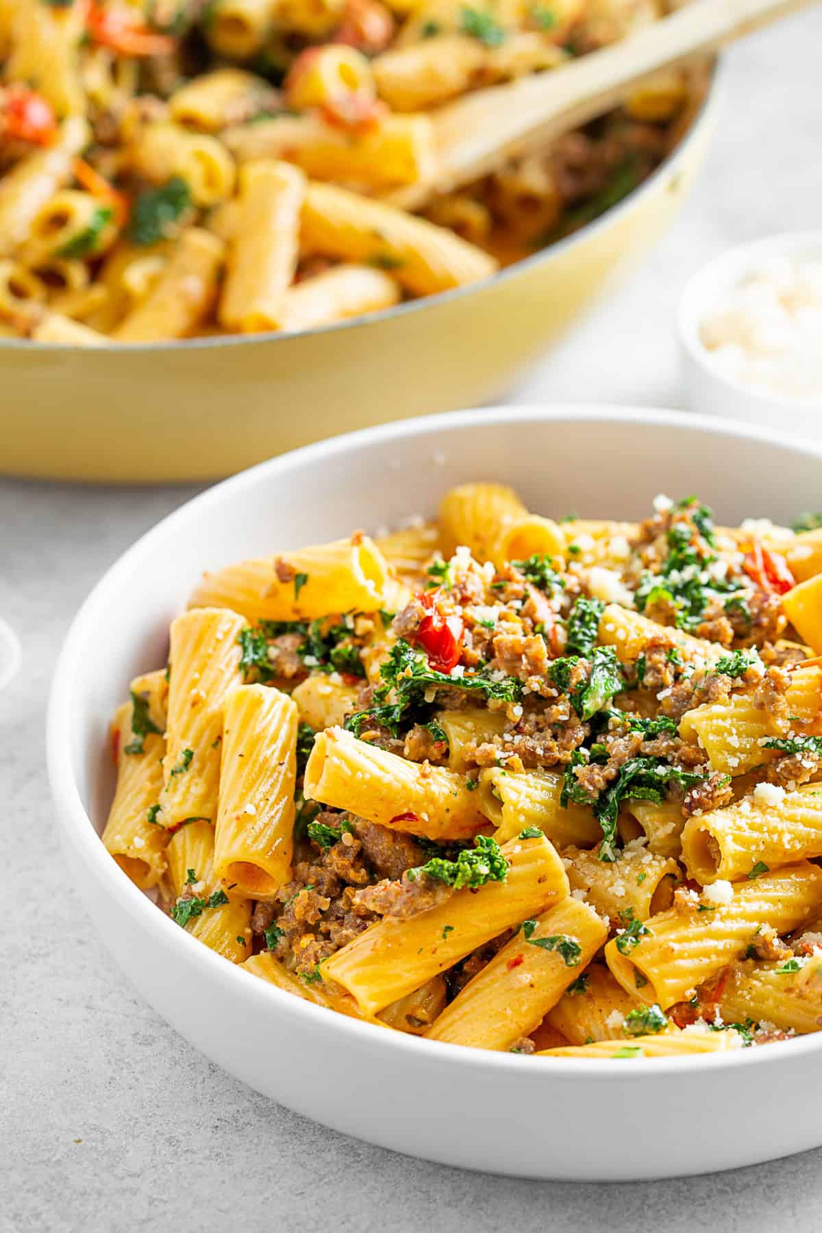 Italian Sausage Kale Pasta in a white bowl with a skillet of pasta in the back. 