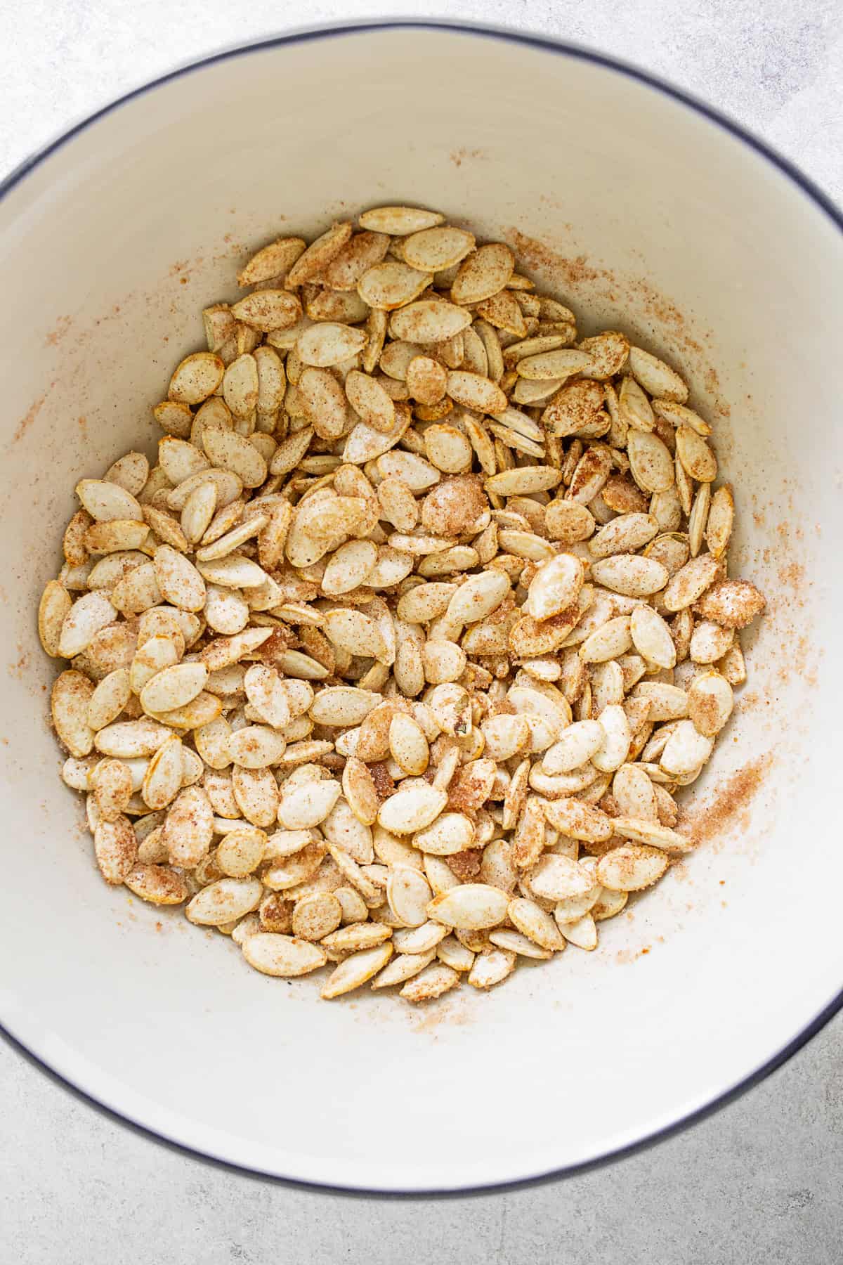 Pumpkin seeds tossed in oil, cinnamon, sugar, and salt in a mixing bowl.