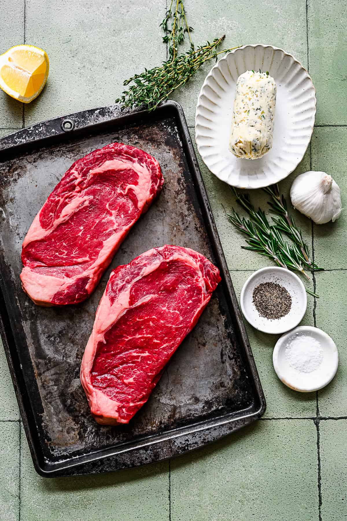 Ribeye steak, seasoning, herbs, and butter on a table. 
