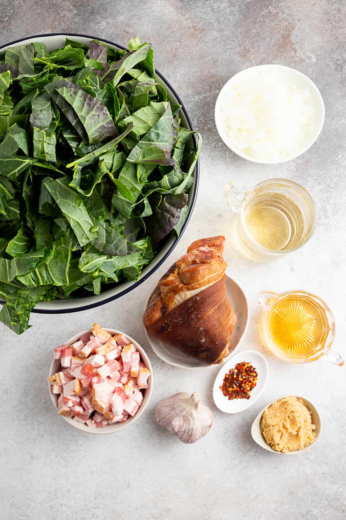 Collard greens, ham hock, bacon, apple cider vinegar, chicken stock, brown sugar, garlic, and red pepper flakes sitting on a table.