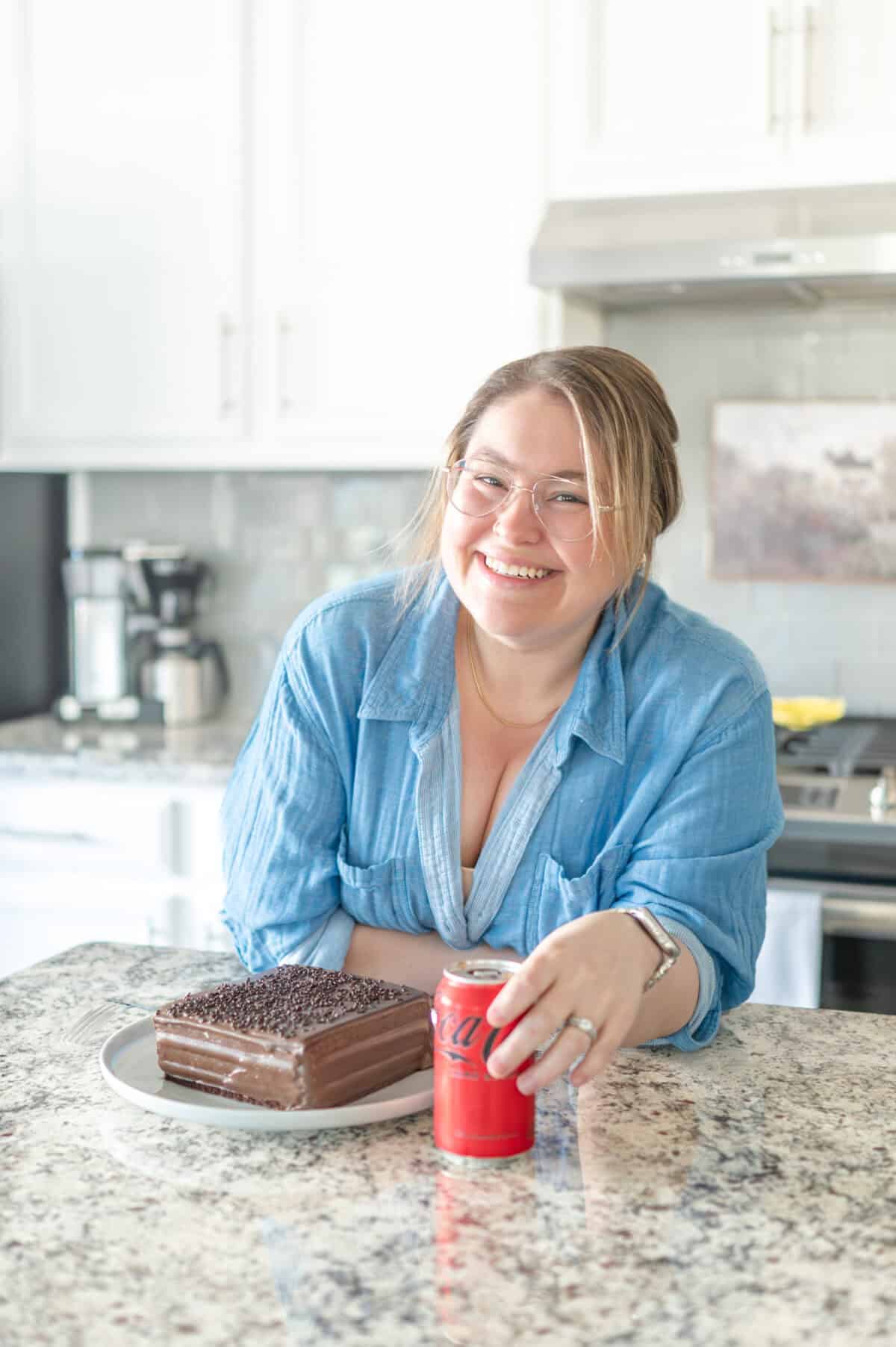 A woman smiling at the camera with a chocolate cake in front of her while she holds a can of coke zero.