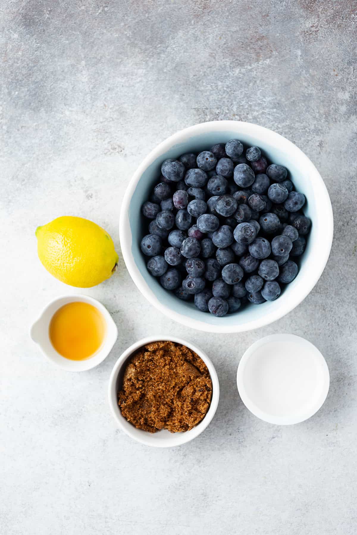 Fresh blueberries, water, a lemon, vanilla extract, and brown sugar sitting on a table.