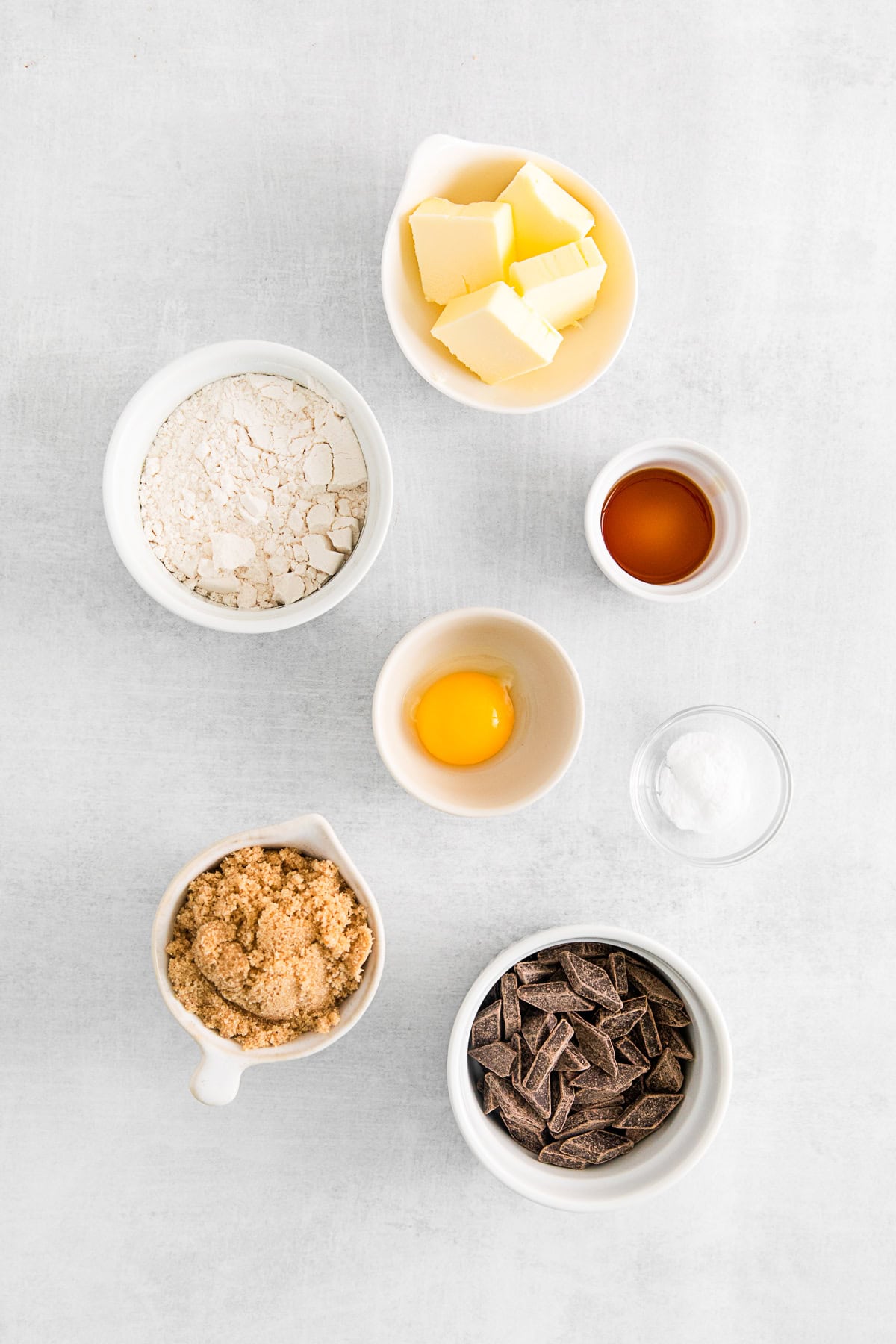 Chocolate chunks, brown sugar, baking soda, egg yolk, vanilla extract, melted butter, and all purpose flour in bowls on a table. 