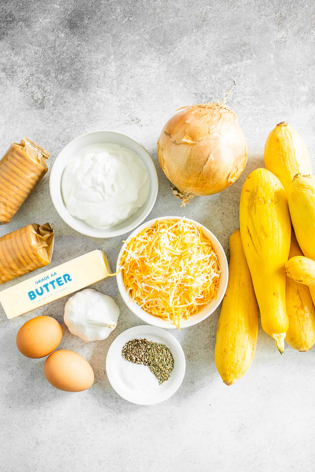 Yellow squash, onion, garlic, eggs, sour cream, cheese, spices, butter, and crackers on a table. 