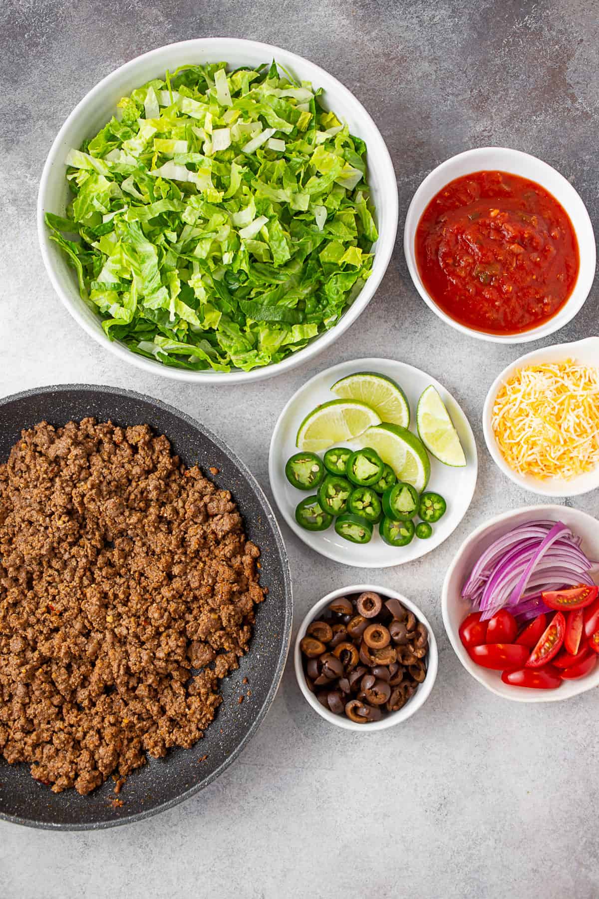 Ground taco meat, lettuce, salsa, cheese, onions, tomatoes, olives, peppers, and limes on a table. 