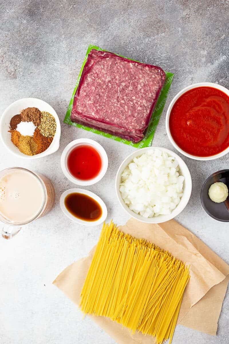 spices, beef stock, tomato sauce, vinegar, onions, pasta, and meat sitting on a table. 